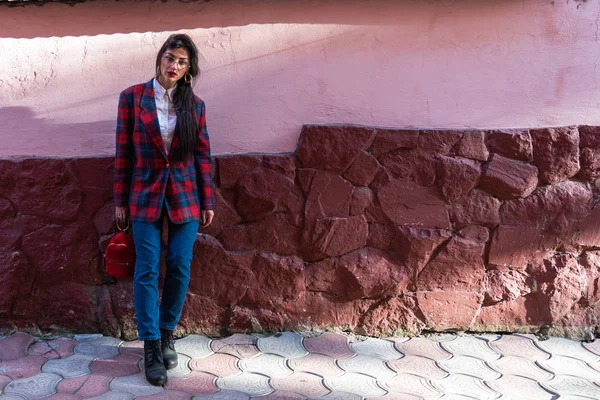 Rua ensolarado foto da bela e elegante jovem mulher de óculos, vestida de jeans azul e jaqueta xadrez, segura pequena mochila vermelha na mão e olhando para a câmera — Fotografia de Stock