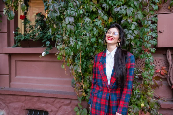 Feliz sorrindo jovem morena feminina em óculos, vestida com jaqueta xadrez e camisa branca, fica perto da parede vermelha com hera verde — Fotografia de Stock