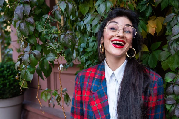 Heureuse femme d'affaires souriante avec des lunettes aux lèvres rouges, vêtue d'une veste à carreaux et d'une chemise blanche, mur rouge avec du lierre vert sur le fond — Photo