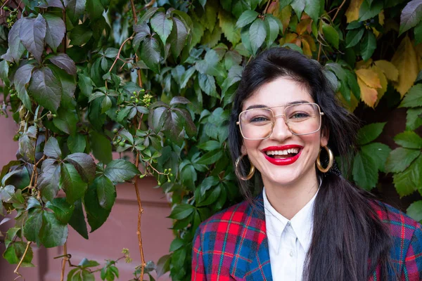 Heureuse femme d'affaires souriante avec des lunettes aux lèvres rouges, vêtue d'une veste à carreaux et d'une chemise blanche, mur rouge avec du lierre vert sur le fond — Photo