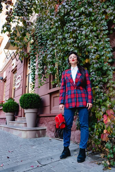 Foto de estilo de rua da linda morena elegante jovem mulher vestida de jeans azul e jaqueta xadrez, segura pequena mochila vermelha e olhando para a câmera, parede vermelha com hera verde no — Fotografia de Stock