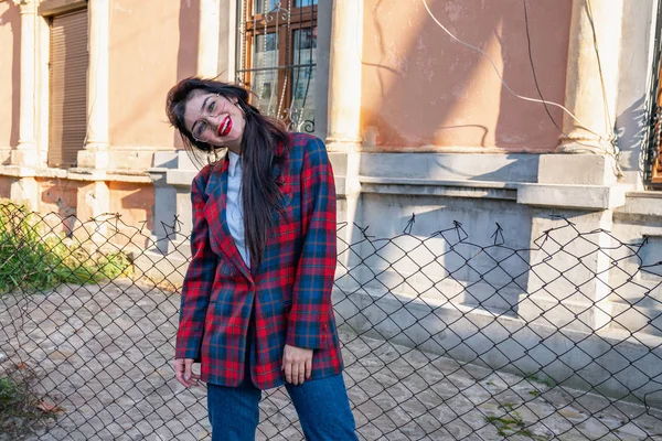 Menina morena Attaractive em óculos vestidos com jaqueta xadrez e camisa branca curvou a cabeça no ombro esquerdo e olhando para a câmera fica perto da casa velha — Fotografia de Stock