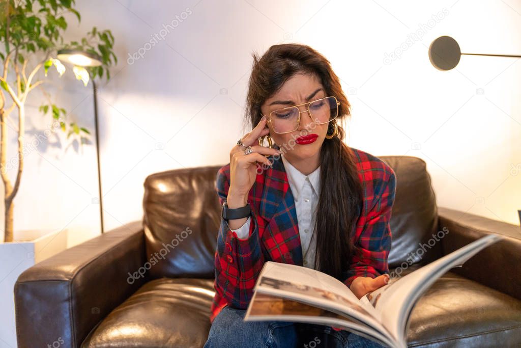 Indignant brunette young woman in plaid jacket sits on the sofa with book in hand