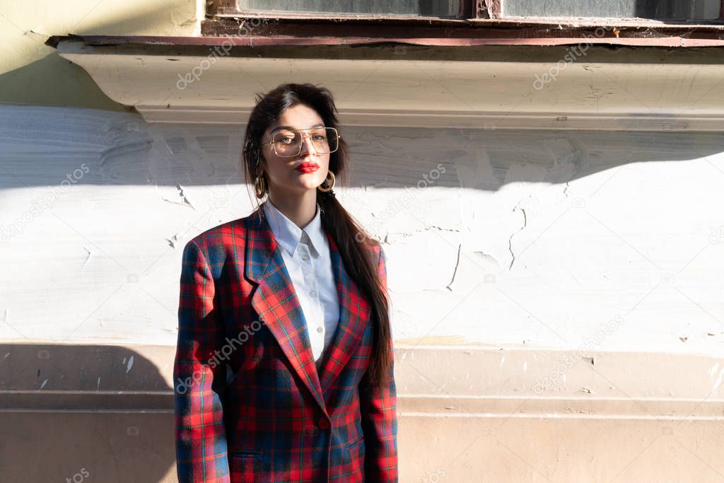 Half lenght portrait of attractive calmy brunette girl in plaid jacket stands in the sun near old wall