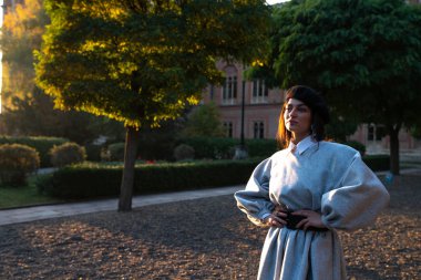 Siyah bereli ve gri elbiseli güzel esmer kız eski tuğladan bir kalenin avlusunda duruyor ve ellerini belinde tutuyor.