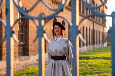 Eski sahte çitin arkasında, siyah bereli güzel bir kız ve gri elbiseli eski tuğla binanın yanındaki yeşil çayır arasında duruyor.