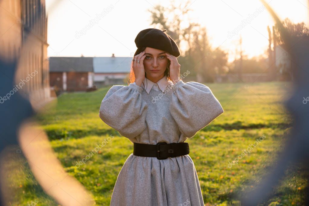 Sad young woman in black beret and grey dress holds her hands near her head, have a headache, stands among the dreen meadow