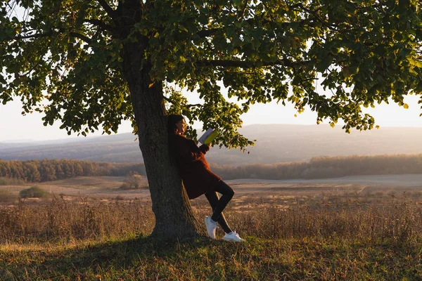Jeune fille adolescente appuyé sur un arbre énorme et tient un livre dans les mains, forêt colorée sur le fond — Photo