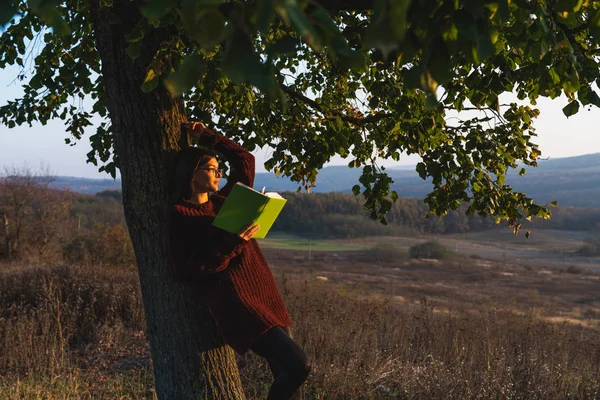 Jolie jeune fille brune en pull rouge appuyé sur un grand arbre, tient le livre à la main et regarde le coucher du soleil — Photo