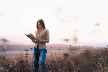 Sıradan pıhtılaşmış gözlüklü güzel esmer kız çayırlarda yürüyor ve arka planda renkli bir orman olan bir kitap okuyor.