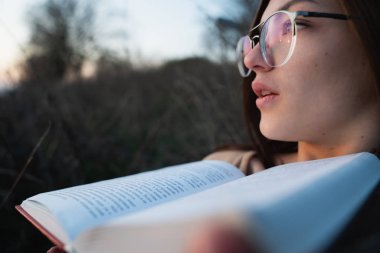 Gözlüklü esmer kızın yakın portresi yüzünün yanında açık bir kitap tutuyor.