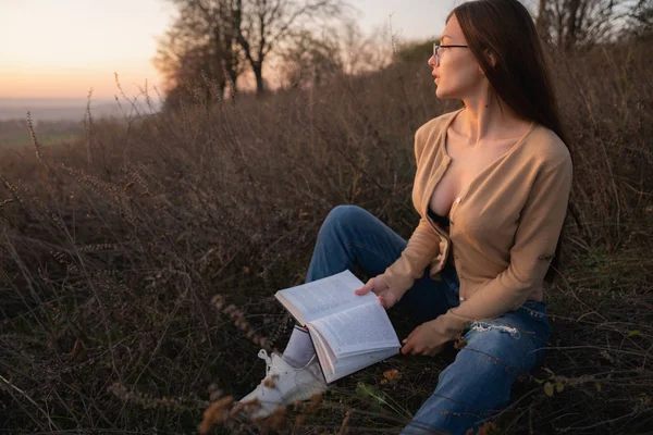 Attractive brunette girl with loose hair dressed in casual clotehs sitting on the groung among the meadow with book in hands and looking away