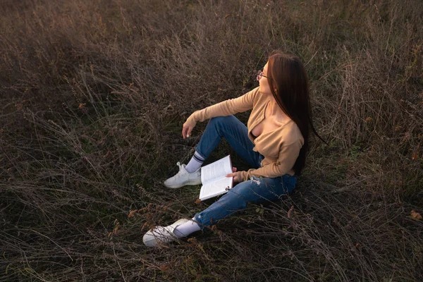 Attractive brunette girl with loose hair dressed in casual clotehs sitting on the groung among the meadow with book in hands and looking away