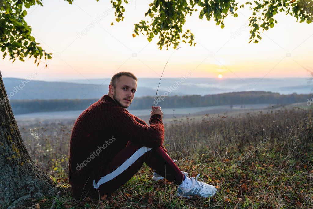 Attractive young man in red sweater sitting under a big tree among the meadow, beautiful landscape on the backgrounf