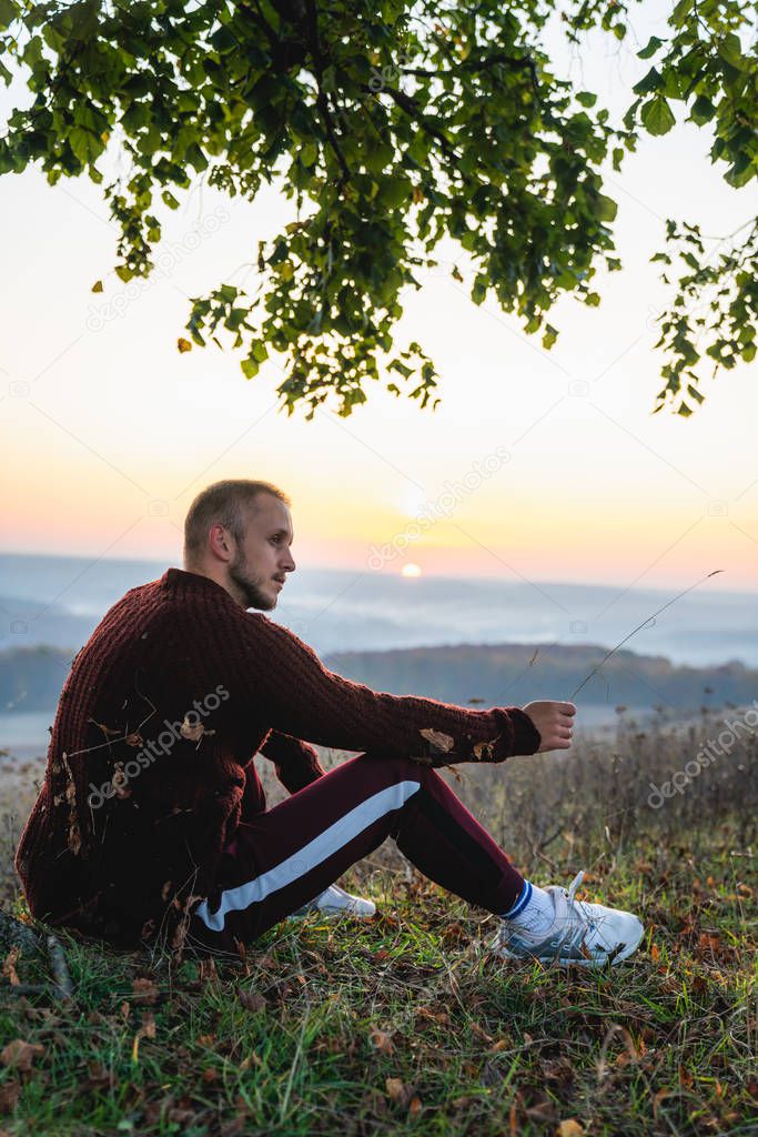 Handsome young man in red sweater sitting on the ground under a big tree, beautiful landscape with sunset on the background