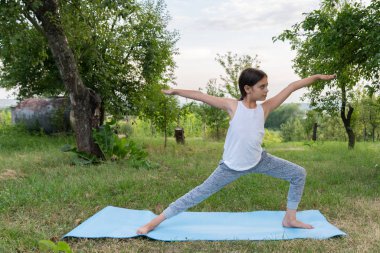 Beyaz fanilalı çekici esmer kız yoga yapıyor, savaşçı iki egzersizi yapıyor, virabhadrasana 2 pozu, parkın ortasında mavi bir minder üzerinde çalışıyor.