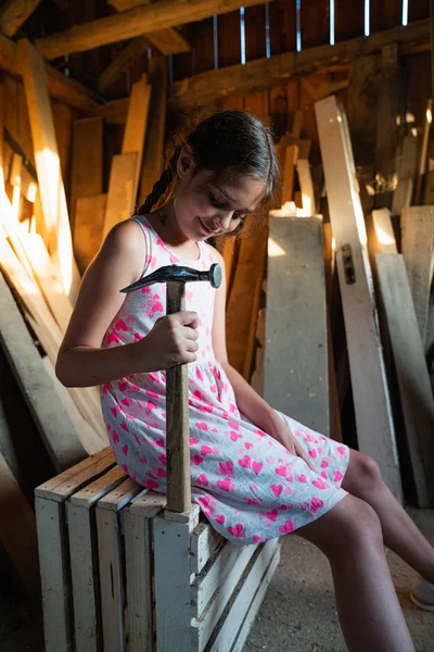 Linda chica morena en vestido blanco y rosa se sienta en la caja de madera entre el taller de carpintería y sostiene el martillo en su mano — Foto de Stock