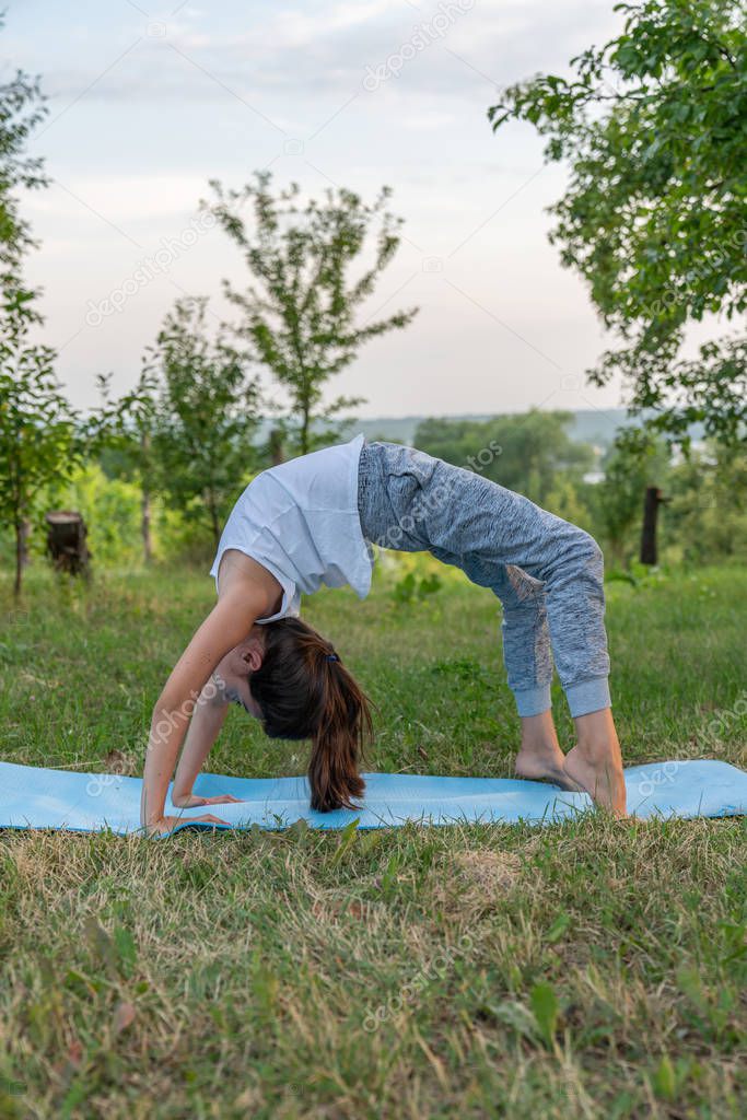 Side view portrait of the young girl in white undershirt doing yoga exercise on the blue mat among the garden, standing in varius poses