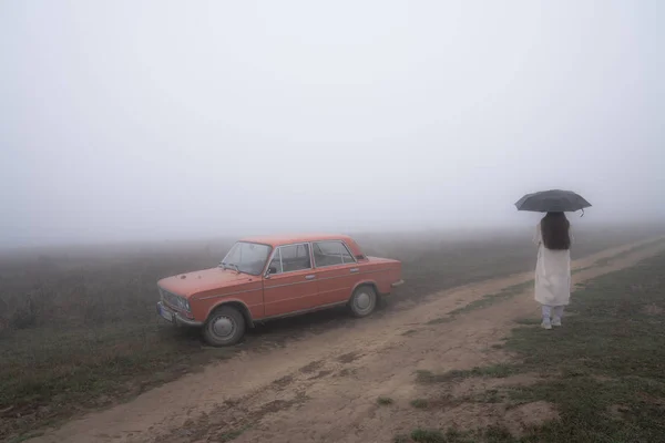 Carro retro vermelho fica perto da estrada de terra no nevoeiro, jovem menina de casaco branco fica sob guarda-chuva — Fotografia de Stock