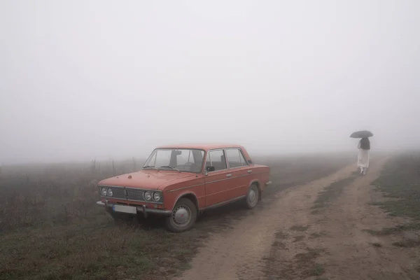 古い赤のレトロな車の汚れの道の近くの霧の中で、白いコートの少女の背景に傘の下を歩くスタンド — ストック写真