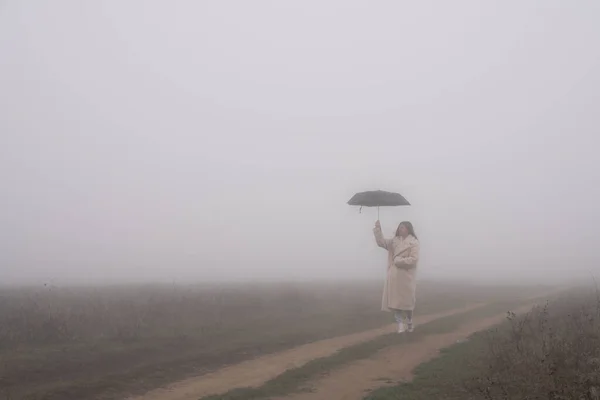 白いコートの若い女の子は未舗装の道路上の牧草地の間で霧の中に立っていると彼女の手に傘を保持 — ストック写真
