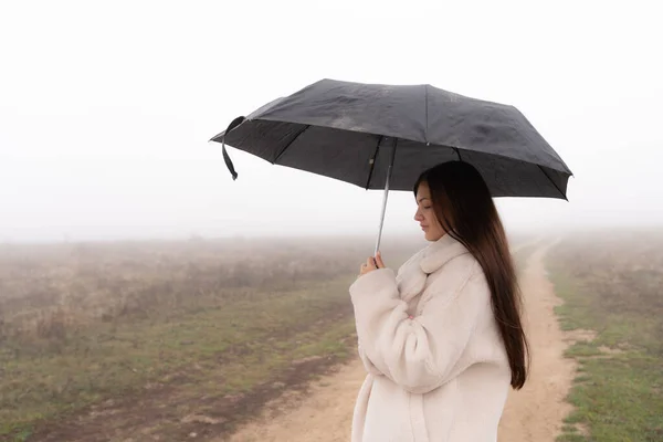 Vita in su ritratto della bella ragazza bruna vestita con cappotto bianco sta sotto l'ombrello, prato nella nebbia sullo sfondo — Foto Stock