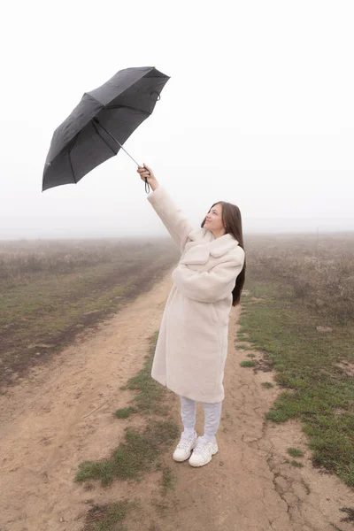 白いコートの魅力的なブルネットの女の子の完全な長さの肖像画は、未舗装の道路の間に立って、右の手で傘を上げ、それを見て — ストック写真