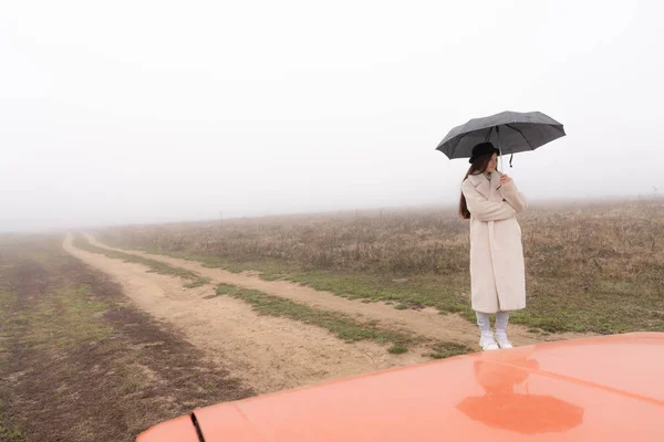 Menina bonita em casaco branco e chapéu preto fica entre a estrada de terra com guarda-chuva na mão, prado no nevoeiro no fundo — Fotografia de Stock