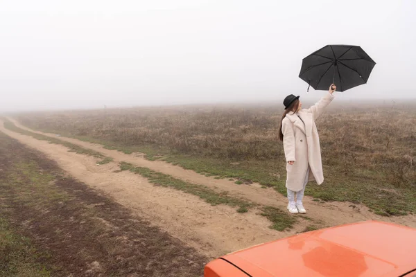 白いコートと黒い帽子の美しい女の子は、手に傘と未舗装の道路の間に立って、背景に霧の中の牧草地 — ストック写真
