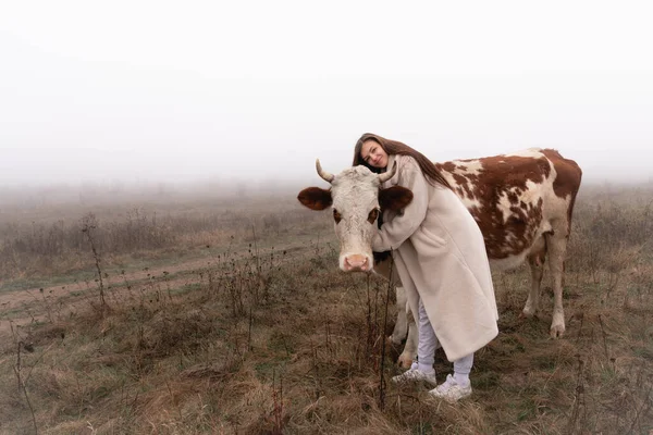 美しいですブルネット女の子で白いコートスタンドの間に霧の中で牧草地で赤と白のcoe 、抱擁彼女とカメラを見て — ストック写真