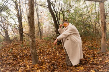 Beyaz ceketli genç esmer kız kütüğe oturup yüzünü portakal yaprağıyla kaplıyor.