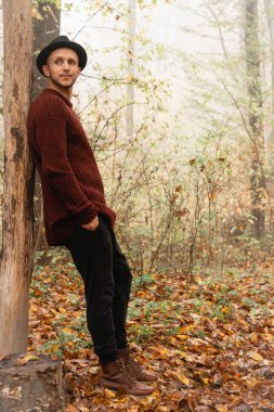 Siyah şapkalı ve kırmızı kazaklı genç adam kesilmiş ağaca yaslanmış ve sisli ormanda el ele tutuşmuş.