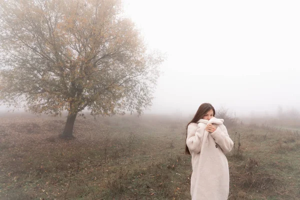 魅力的なブルネットの女の子は白いコートに包まれた草原の間に立つ、背景に霧の中で大きな木 — ストック写真