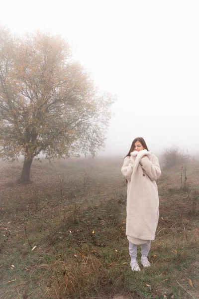 魅力的なブルネットの女の子は白いコートに包まれた草原の間に立つ、背景に霧の中で大きな木 — ストック写真