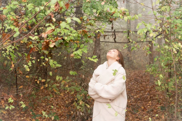 Chica morena feliz con abrigo blanco se levanta con las manos cruzadas entre el bosque en la niebla y mirando hacia arriba — Foto de Stock