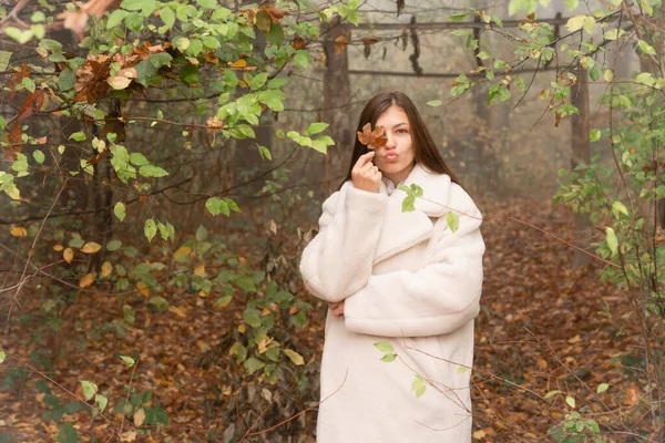 Muchacha morena atractiva en abrigo blanco se encuentra entre el bosque en la niebla, cubierto un ojo con hoja de naranja y mirando a la cámara — Foto de Stock