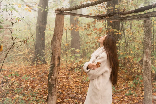 Young girl with brunette loose hair dressed in white coat stands among the forest in fog with crossed hands and looking up