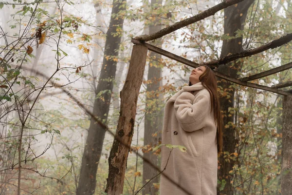 Menina morena atraente envolto em branco casaco fica entre a floresta no nevoeiro — Fotografia de Stock