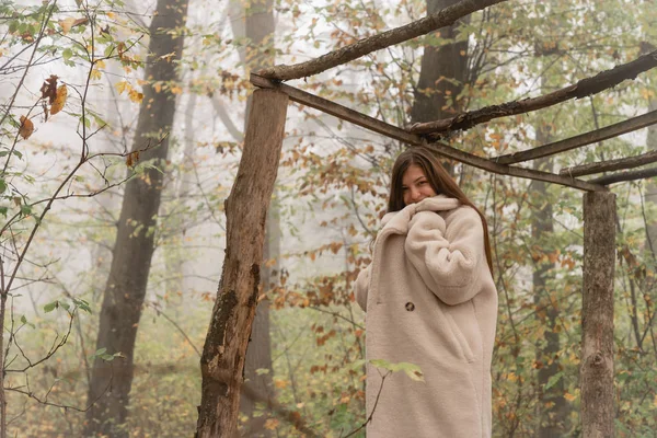 Menina morena atraente envolto em branco casaco fica entre a floresta no nevoeiro — Fotografia de Stock