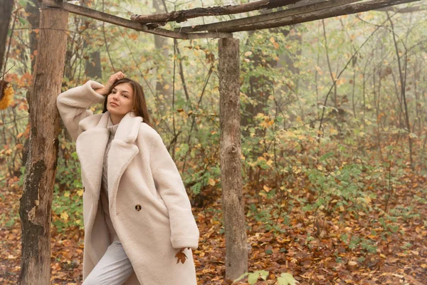 Hermosa morena de abrigo blanco apoyada en un árbol caído entre el bosque en la niebla y mirando a la cámara — Foto de Stock