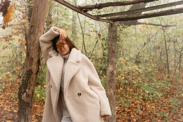 Hermosa morena joven con abrigo blanco apoyada en un árbol caído entre el bosque de niebla y cubriéndose la cara con hojas de naranja — Foto de Stock