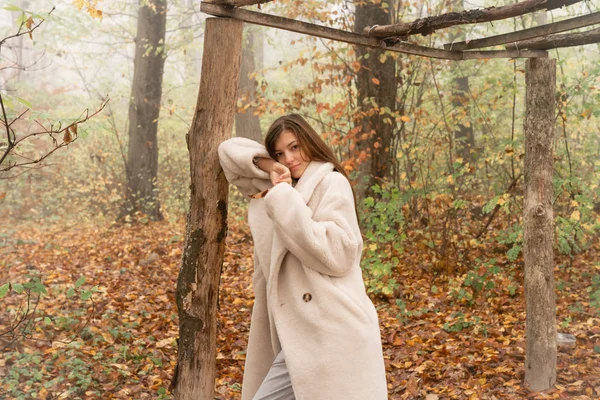 Piękna brunetka w białym płaszczu pochylona na ściętym drzewie pośród lasu we mgle i patrząca w kamerę — Zdjęcie stockowe