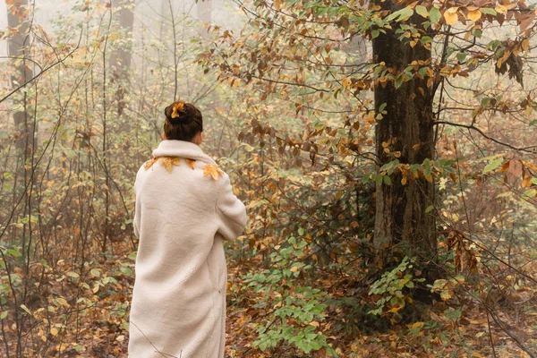 Morena joven mujer vestida con capa blanca esparcida con hojas amarillas se aleja a la cámara entre el bosque en la niebla — Foto de Stock