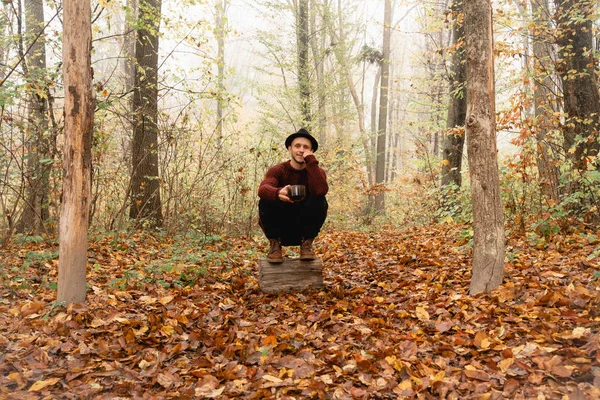 Hombre joven con sombrero negro y suéter rojo agachado en el muñón entre el colorido bosque, sostiene la copa en su mano y mirando a la cámara — Foto de Stock