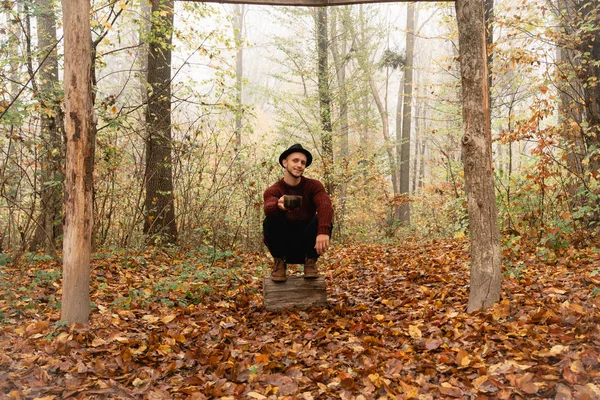 Hombre joven con sombrero negro y suéter rojo agachado en el muñón entre el colorido bosque, sostiene la copa en su mano y mirando a la cámara — Foto de Stock