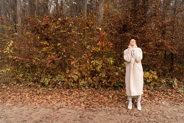 Hermosa chica morena envuelta en abrigo blanco se encuentra entre el colorido bosque con los ojos cerrados, copsoacefor su texto — Foto de Stock