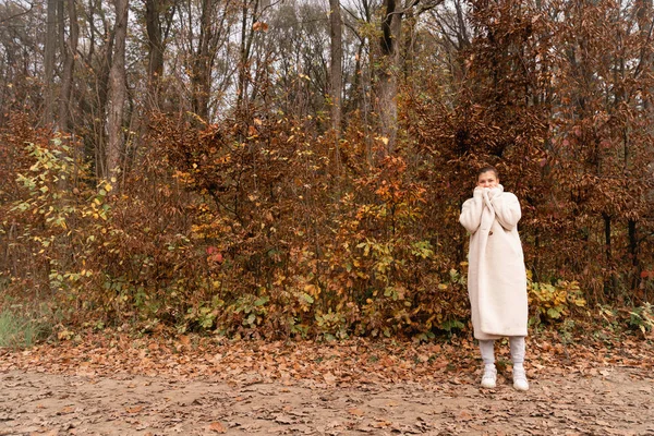 Atraente morena jovem fêmea envolto em branco casaco fica entre a floresta colorida com olhos fechados — Fotografia de Stock