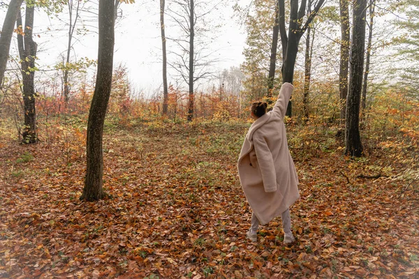 Joven morena hembra en abrigo blanco se para con la mano levantada de nuevo a la cámara entre el colorido bosque — Foto de Stock