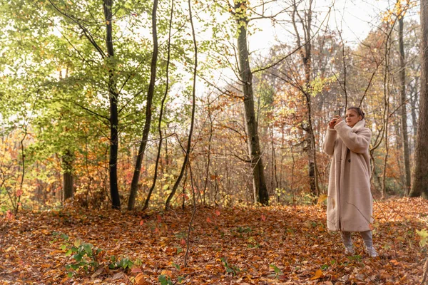 Atractiva hermosa chica morena en abrigo blanco caminando entre el colorido bosque y haciendo fotos en su teléfono móvil — Foto de Stock