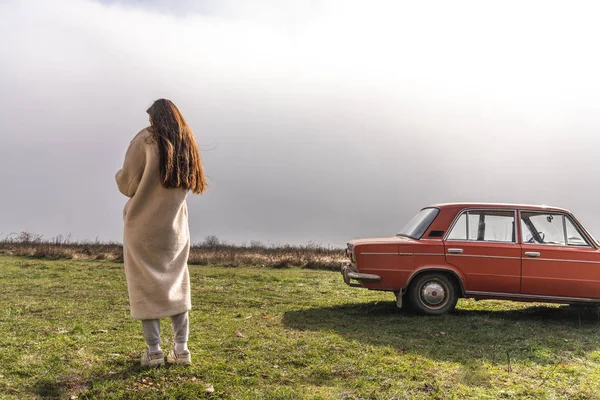 Joven morena hembra con abrigo blanco se para al sol de nuevo a la cámara cerca del coche retro rojo, nubes grises en el fondo — Foto de Stock